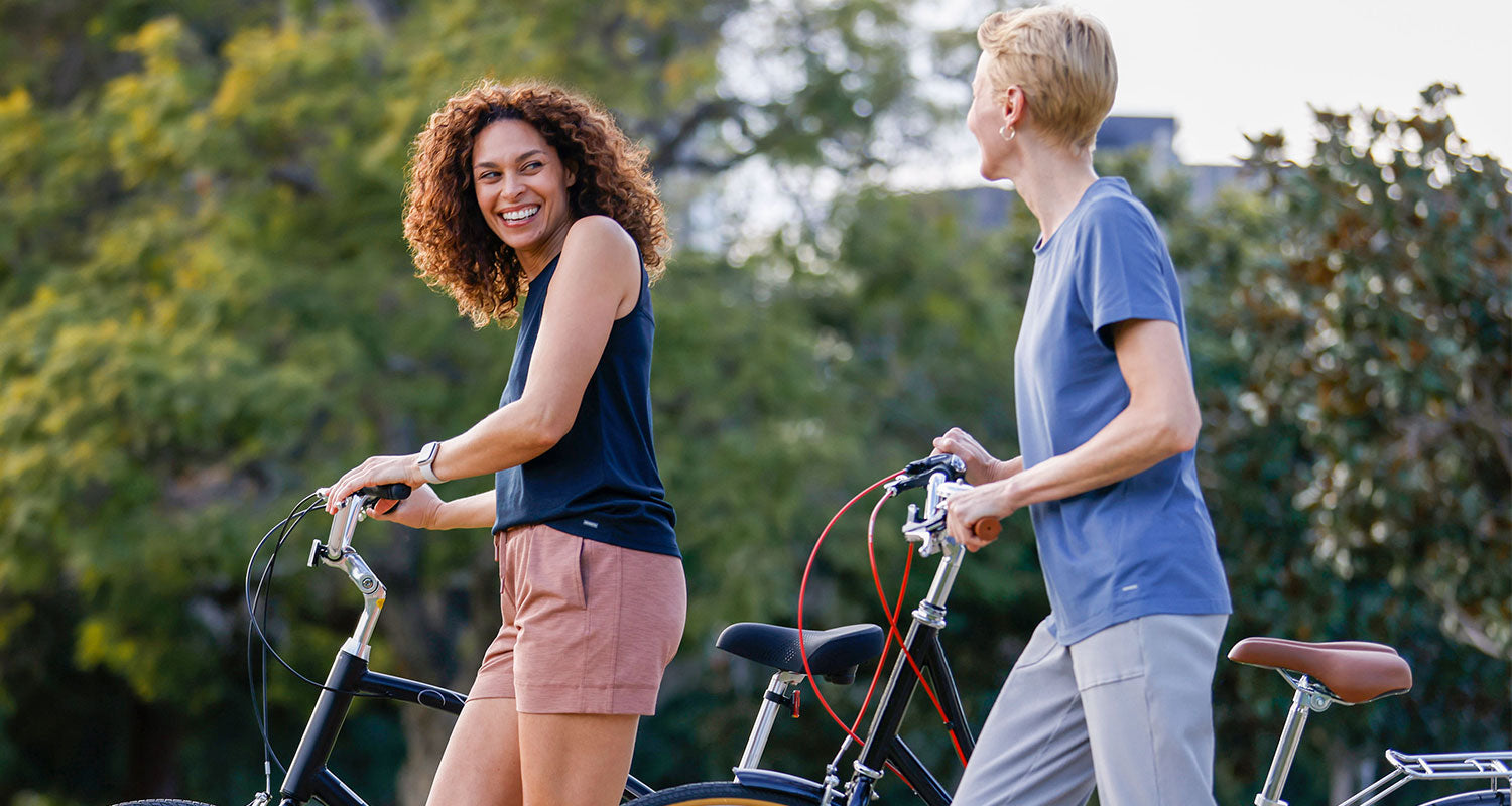 Two female friends laughing and walking their bikes through a park